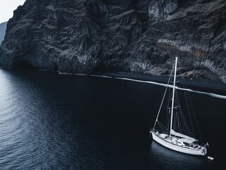 Aerial view from the side of a small sailboat with one mast, sails down, anchored near a volcanic black sand beach with dramatic cliffs in the background