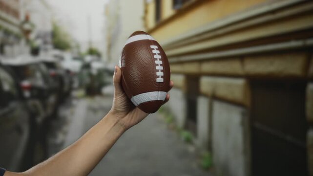 Man holding football while standing outdoors on a city street, captured in an urban setting with a focus on sports and casual outdoor activity.