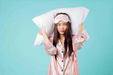Stressed Asian woman with pillow covers her ears because of snore, pajama and sleep mask show her...