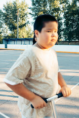 boy riding black and green kick scooter on outdoor urban court in summer evening, beige t-shirt and grey shorts playful childhood moment healthy lifestyle, street activity, motion blur