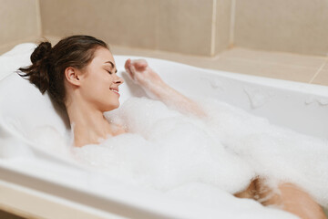 Relaxed young woman enjoying a bubble bath in a modern bathroom, smiling with contentment as she...