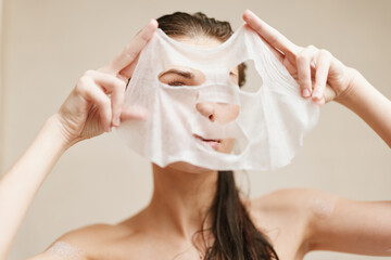 Young woman enjoying skincare routine while applying facial mask in a bright bathroom, emphasizing...