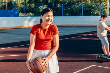 Asian teen smiling girl enjoys basketball game on urban court with boy on scooter in background