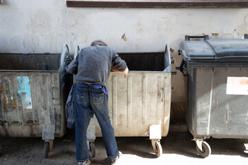 Individual rummages through dumpsters in a city alley, searching for food or discarded items in the sunlight