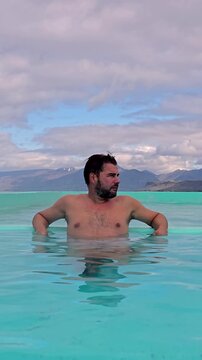 A man enjoys the soothing geothermal waters of Icelands Westfjords, surrounded by dramatic landscapes. The day is calm with gentle clouds and striking mountain views in the distance.