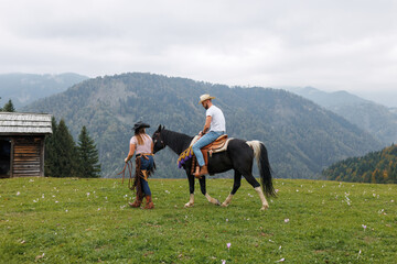 A man in cowboy attire rides a horse with assistance from a woman in a rural area of Styria,...