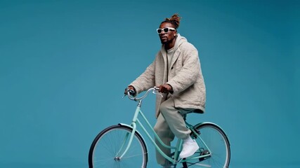 A stylish african american man in sunglasses rides a bicycle against a blue background, a concept for urban fashion, sustainable lifestyle, and modern trends