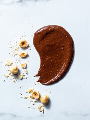 Close-up of creamy chocolate hazelnut spread on a white marble background, styled with whole and crushed hazelnuts. The rich, smooth texture of the spread contrasts beautifully with the crunchy nuts, 