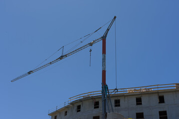 A large construction crane with a long boom stands over a building under construction, set against a clear blue sky on a sunny day.
