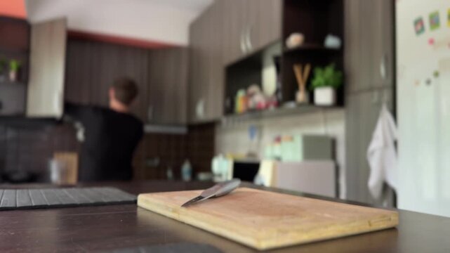 A frustrated man frantically searches kitchen shelves, then storms out in anger and forcefully stabs a knife into a cutting board. Emotional indoor scene, home setting.