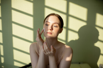 Radiant Woman Applying Skincare in Soft Morning Light with Shadow Patterns on Green Wall
