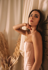 Fototapeta premium A woman in a pink satin corset posing near dried flowers and a brick wall. 