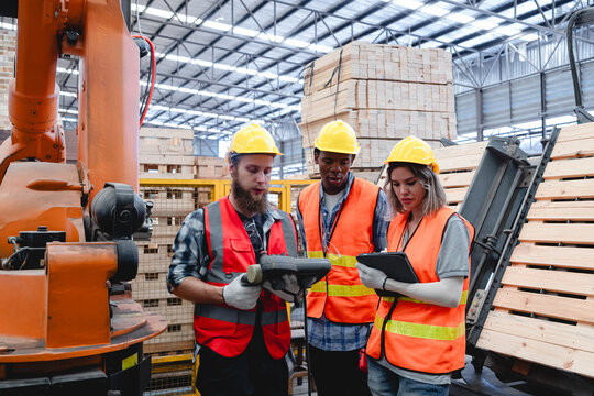 Diverse team of industrial workers wearing helmets and safety vests standing near robotic arm, reviewing data on digital tablets inside smart pallet manufacturing facility. The scene reflects teamwork