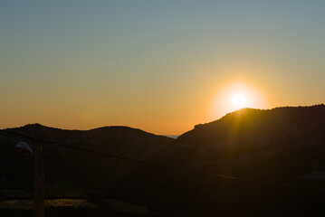 The bright sun sets behind a silhouetted mountain range, casting a warm orange and yellow glow across the sky with soft blue at the top.