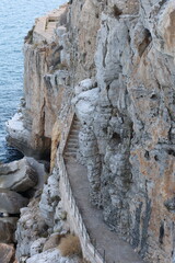 A narrow stone path winds along a steep seaside cliff, partially carved into the rugged rock face. The dramatic walkway offers breathtaking views of the sea below and the textured cliffs.