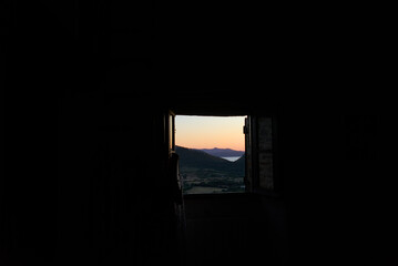 A vibrant sunset over a landscape of hills and water is visible through a dark, open window frame, creating a striking contrast.
