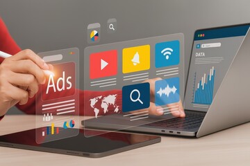 A person interacts with digital marketing and data analytics icons on transparent screens beside a laptop and tablet on a desk.