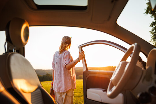 Joyful woman stepping out of car into golden sunset light, capturing feeling of carefree travel during summer road trip. View from car interior