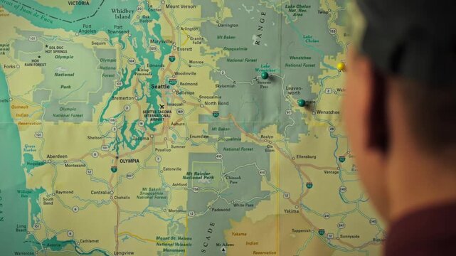A person examines a map of Washington state, pointing to various locations while planning a hiking adventure. The focus is on outdoor exploration and route selection
