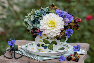 Blumen-Arrangement mit weißer Dahlie, Federnelken, Hortensienblüten und Brombeerzweigen in einer Porzellantasse