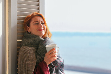 Ginger-haired woman, 30s, standing   by the window in cozy covered in blanket, reading drinking coffee. Calm, peaceful moment in soft natural light.