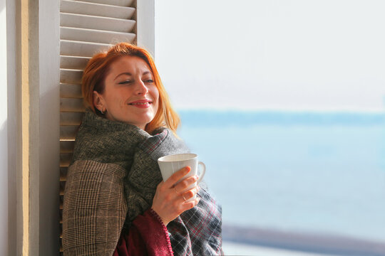 Ginger-haired woman, 30s, standing by the window in cozy covered in blanket, reading drinking coffee. Calm, peaceful moment in soft natural light. - Powered by Adobe