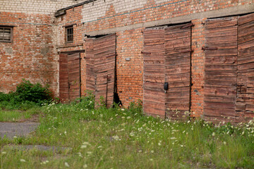 The photograph shows an old brick building with several wooden, slanted doors. In front of it is an area overgrown with grass and weeds.