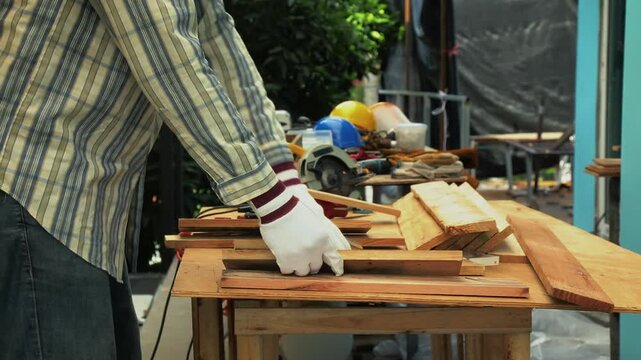 Carpenter cutting wooden plank with circular saw on outdoor bench while wearing gloves and protective mask
