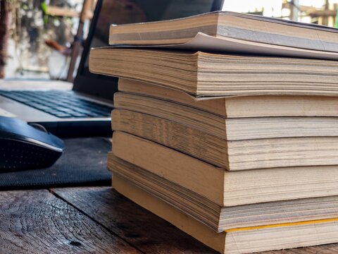 A stack of softcover books on a wooden table beside a mouse and laptop in natural sunlight. Warm tones, visible paper texture, shot from a low angle. Suitable for study or reading themes.