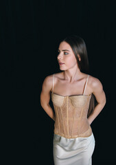 A young woman wearing a beige lace corset and white satin skirt poses gracefully against a black...