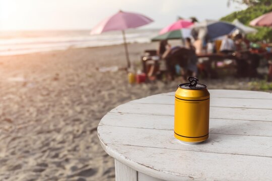 Open yellow aluminum can stands on a white wooden table beach and people under umbrellas in the blurred background
