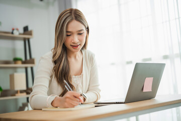 Young woman engaged in creative work at home, writing notes in a cozy workspace with laptop,...
