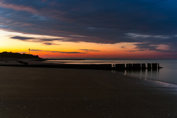 Sunrise on the beach near the Punta Tagliamento lighthouse in Bibione, Italy, with a wooden breakwater in the foreground and calm sea under colorful sky.