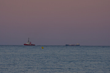 A red and white tugboat is visible on the calm, blue ocean under a soft, pastel sky during dusk or dawn, with yellow buoys floating in the foreground.