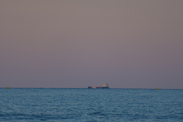 Fototapeta premium A distant cargo ship is seen on the calm blue ocean under a soft, pastel-colored sky during dusk or dawn, with a hint of a yellow buoy in the far right.