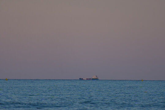 A distant cargo ship is seen on the calm blue ocean under a soft, pastel-colored sky during dusk or dawn, with a hint of a yellow buoy in the far right.