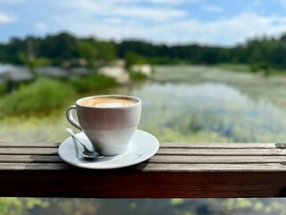 Cup of coffee on a wooden railing with a peaceful lake and forest landscape in the background on a sunny day