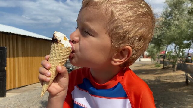 A young boy takes a big bite of a chocolate-dipped ice cream cone. The sunny outdoor setting in Novofyodorovka, Crimea adds to the joyful summer moment.