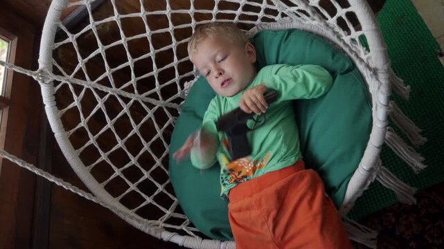 A young boy in green and orange pajamas in a white, woven hanging chair with a large green cushion. He is dangerously holding a black handgun in his hand