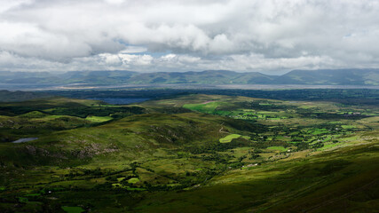 Fototapeta premium Rolling green valleys with interspersed forests and open pasture lead to distant mountains under a cloudy sky, taken from Coomloughra Horseshoe scenic landscape in Ireland.