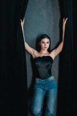 A confident young woman poses wearing a black corset and blue jeans.
