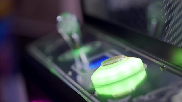 A brightly lit button on a claw machine is being pressed, signaling the start of a game. The joystick and control panel are visible in a lively indoor arcade environment.