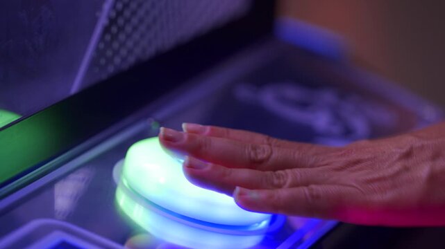 A close-up of a finger pressing a glowing red and green button on a claw machine control panel. The vibrant arcade environment surrounds the game, heightening excitement and focus.