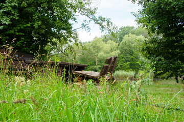 Im Vordergrund frisches, h&ouml;heres Gras. Dahinter ein h&ouml;lzerner Rastplatz mit Tisch und Bank. Eine herausstehende Schraube macht die Bank weniger einladend.
