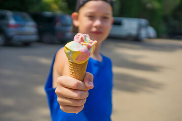 A young boy wearing a blue t-shirt and a cap, holding out a colorful ice cream cone towards the camera, with blurred background, a sunny outdoor setting, concept of summer treats, childhood joy