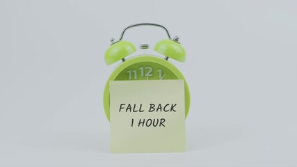 Fall Back 1 Hour word written on a sticky note placed in front of a green ringing clock on white background, symbolizing daylight saving time, clock adjustment, and schedule change concept