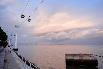 A scenic view of a waterfront promenade with a cable car system overhead, trees lining the walkway, and a calm sea under a cloudy sky. © luengo_ua