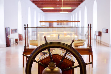 A maritime museum interior featuring a ship's wheel in the foreground and ship models in glass cases in the background.