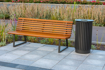 Modern wooden bench sits beside black waste bin on concrete patio with ornamental grasses creating functional urban seating area