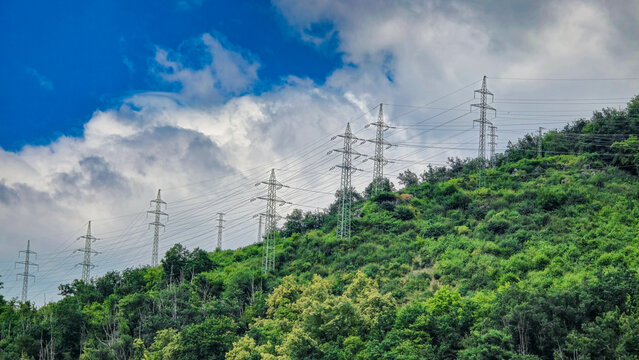 Power lines on a green hill with dense vegetation and cloudy sky. Concept of energy, infrastructure and power transmission.